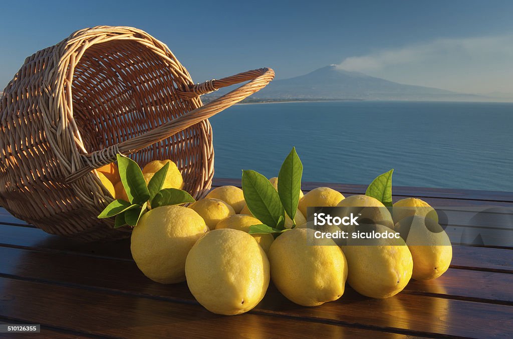 Dreaming of Sicily Wicker basket full of  lemons on a wooden table with blue sea and mount Etna in the background Lemon - Fruit Stock Photo