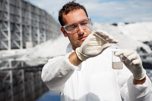 Scientist in white safety suit examing polluted water in a river at industrial site.
