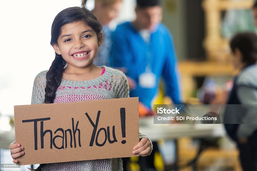 Pretty Hispanic girl holds 'Thank You!' sign in soup kitchen Cute Hispanic girl is holding a cardboard sign with 'Thank You!' witten on the board. She and her family are in a soup kitchen or food bank. She is smiling at the camera. Her brown hair is in a braid. Volunteers ar serving her family in the background. Focus is on the girl and the sign. Child Stock Photo