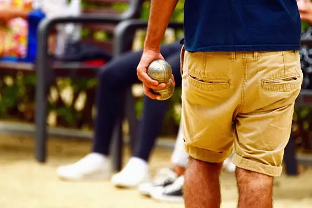 People playing boules during summer hollidays. There is a man close-up, the rest of players waiting. The scene is in a park with sand.