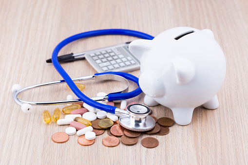 White piggy bank on a wooden desk with coins, medicine on the side and a calculator in the background with a stethoscope on top