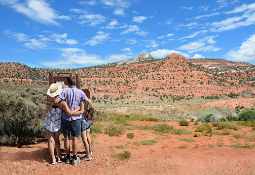 Father and daughters hugging and looking at map for direction on their hiking trip. Cloudy sky and red mountains in the background. Girl is wearing backpack. New Mexico, USA.