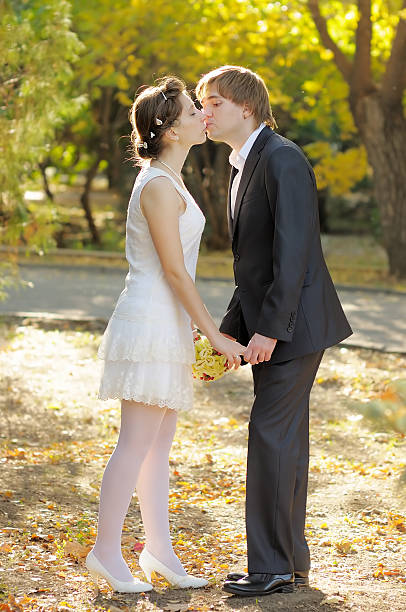 Just married couple kissing each other on the roud in the middle of the forest at summer. She has a beautiful wedding dress with big veil and he is wearing elegant suit