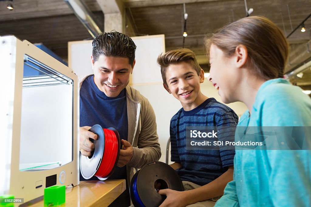 Children learning how to use 3D printers in makerspace studio Mid adult Hispanic man is teaching his children how to use a 3D printer in a makerspace studio. Father and preteen son are holding rolls of colorful filament. Excited elementary age little girl is watching cube style 3D printer create a 3D plastic printed object. 3D Printing Stock Photo