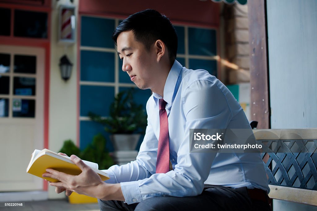 Asian Man Reading a Book Sitting on Bench Asian Man in Business Suit Reading a Book Sitting on Bench Adult Stock Photo