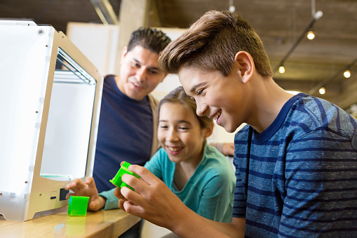Preteen and elementary age siblings are using a 3D printer with their father in a makerspace studio. Family is printing 3D plastic objects with a cube style 3D printer. Mid adult man is watching his children learn about modern technology.
