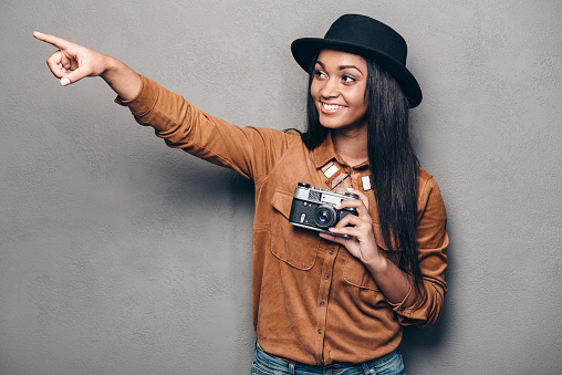 Beautiful cheerful young African woman holding retro styled camera and pointing away with smile while standing against grey background