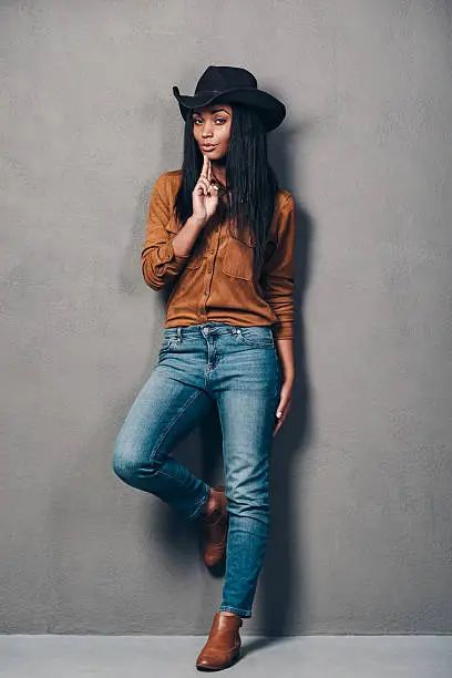 Full length of beautiful young African woman in hat gesturing handgun and looking at camera while standing against grey background