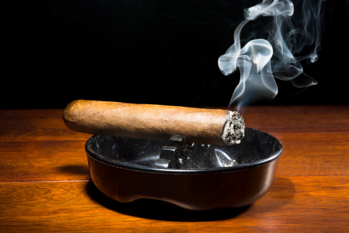 A burning cigar in a classic black ashtray streaming smoking in a dark, moody setting. The smoke is real, straight from the cigar and not put in later during post processing.