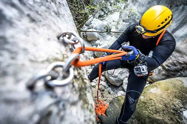 Member of canyoning team with camera attached to his arm is moving down the cliff in the canyon. The rope is secured with special alpine system.