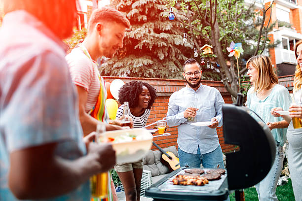 Young People Having Fun At Barbecue Party. Group of young people having fun at barbecue party in a backyard. Man with apron grilling and giving meats to his friends. African man holding salad. They are enjoy spending time together. grill burgers stock pictures, royalty-free photos & images