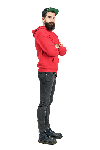 Side view of young bearded man in red hoodie wearing baseball cap with crossed arms. Full body length portrait isolated over white studio background