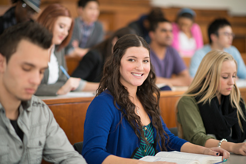 A multi-ethnic group of college age students are in class listening to their professor give a lecture. One woman is smiling while looking at the camera.