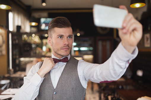 Elegant young man fixing his bow tie and taking a selfie at an exclusive gentlemen's bar.