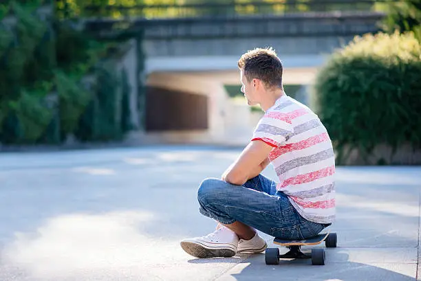 A side photo of a young man sitting on his skateboard and waiting
