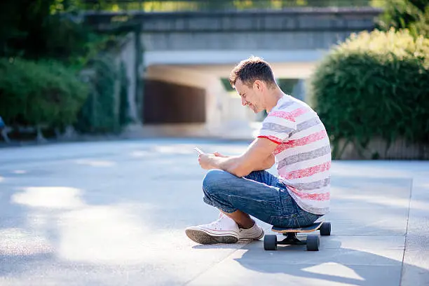 A side photo of a young man sitting on his skateboard and texting