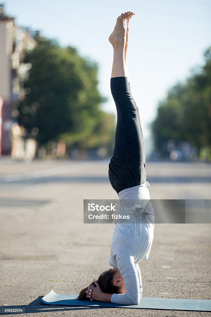 Street yoga: salamba sirsasana pose Yoga in the city: beautiful young sporty woman working out on the road on summer day, doing supported headstand posture, salamba sirshasana, full length, profile view Abdominal Muscle Stock Photo