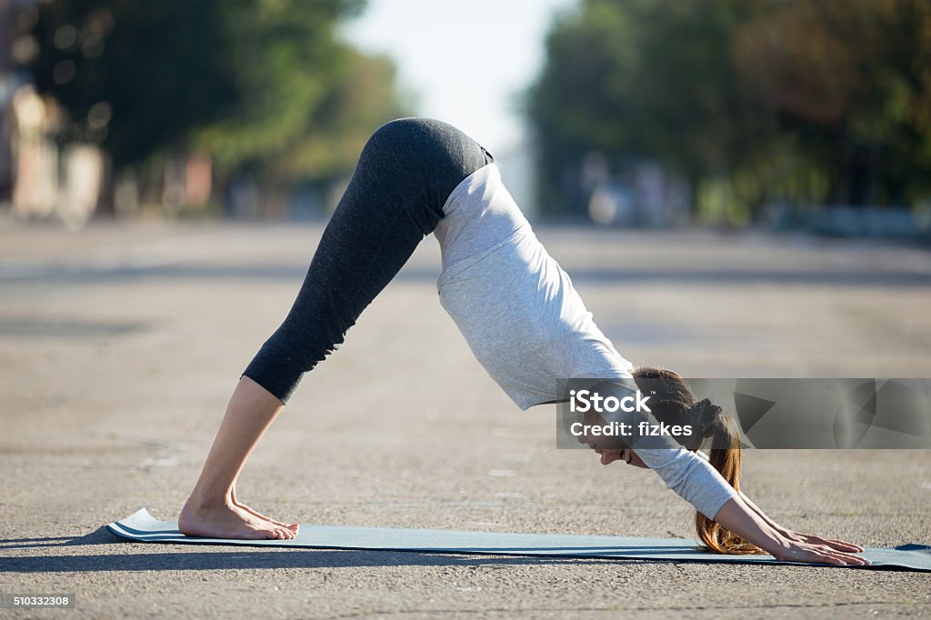 Street yoga: Downward facing dog pose Yoga in the city: beautiful young sporty woman wearing sportswear working out on the street on summer day, doing downward facing dog posture, full length, profile view Downward Facing Dog Position Stock Photo