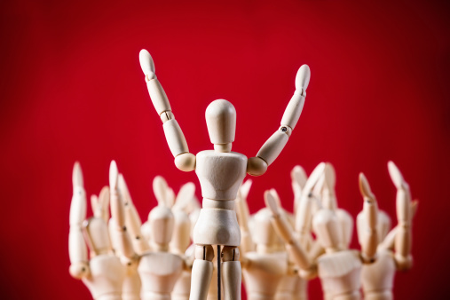 A little wooden figurine addresses an out-of-focus collection of other wooden puppets, all with their arms raised, at a rally, meeting, seminar, exercise class or theatrical performance.Red background with copy space.
