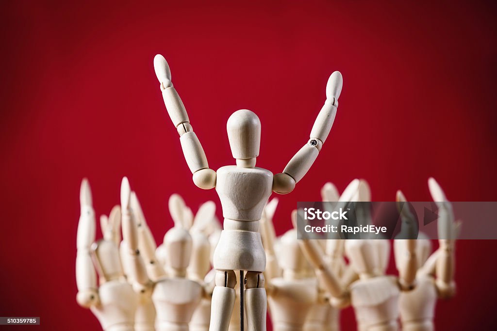 All together now! Puppet faces others, all with raised hands A little wooden figurine addresses an out-of-focus collection of other wooden puppets, all with their arms raised, at a rally, meeting, seminar, exercise class or theatrical performance.Red background with copy space. Populism Stock Photo