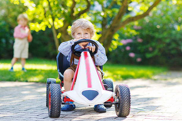 Active little boy driving pedal car in summer garden Active little boy of 3 years driving pedal car in summer garden, outdoors. His little brother on background. kid toy car stock pictures, royalty-free photos & images