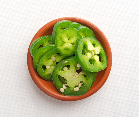 Top view of a small bowl of sliced jalapeno peppers on an off white tablecloth illuminated with natural light.