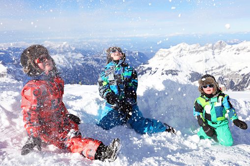 Three cheerful kids are having fun and laughing while playing in the snow on a nice sunny winter day in the mountains in Switzerland. The boys are dressed in colourful ski clothing in green, red, and blue colors. This image is taken on top of Mount Titlis (10,000ft) in Engelberg in Swiss Alps.
