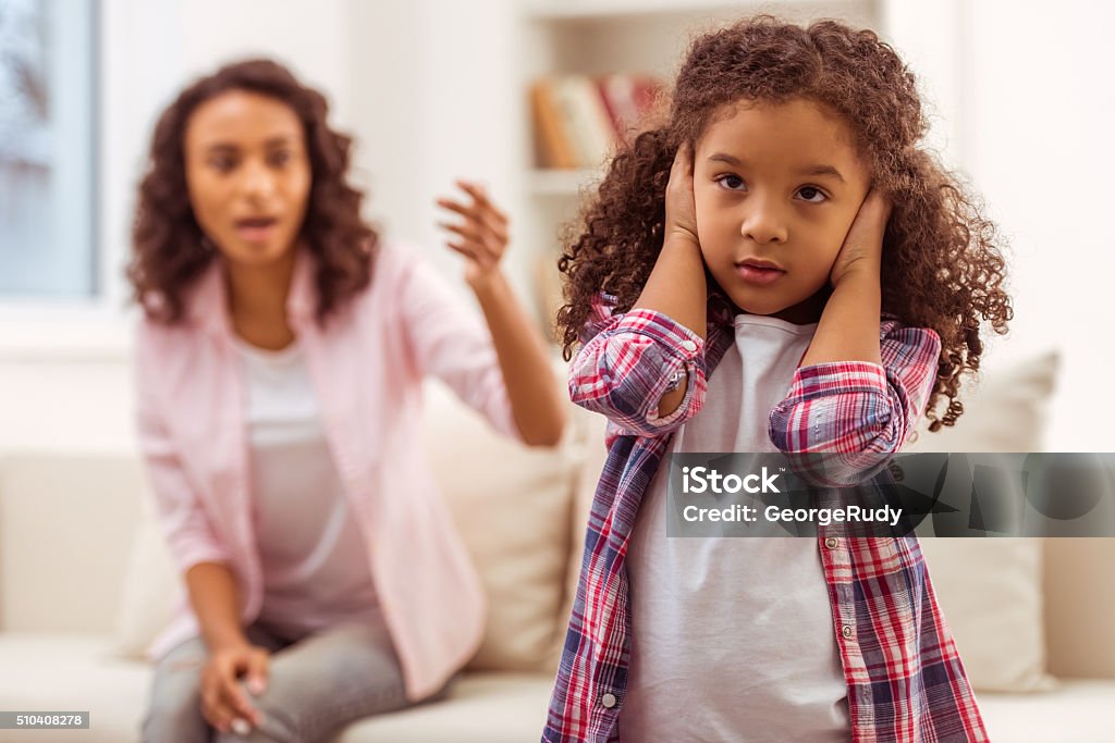 Mother and daughter Cute little Afro-American girl covering her ears, in the backgroung mother scolding her. Child Stock Photo