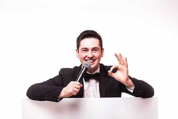 Young elegant talking man holding microphone talking on table  with hands sign. Isolated on white background. Showman concept.