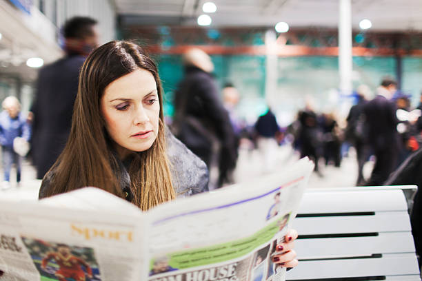 One young woman sits in a train station reading a newspaper while other passengers walk behind her. Made in 2013 London's lypse.
