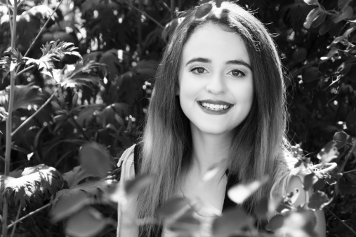 Horizontal B&W outdoor garden shot of smiling teen girl with delphiniums in background.