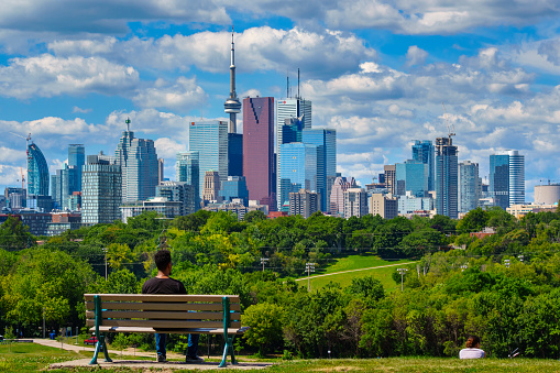 Toronto, Canada - July 22, 2015: Young man sits on a bench in Riverdale Park and looks at the view with the skyline of Toronto in the background.
