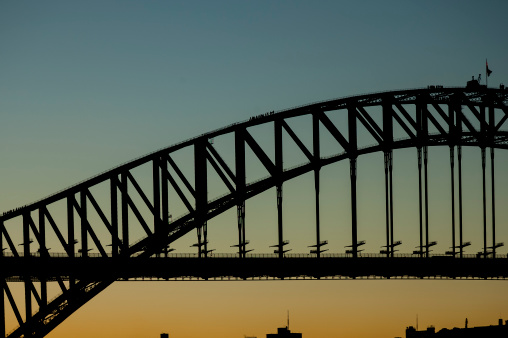 Sun setting with a clear sky behind Sydney Harbour Bridge. Silhouette of half of the bridge with groups of climbers captured on their way to the top.