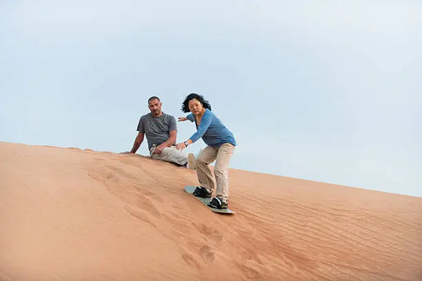 Dubai iStockalypse.  A  Middle Eastern sand boarding instructor watches a mature female adventure tourist as she learns to sandsurf down a dune in the red desert between Dubai and Sharjah, United Arab Emirates.