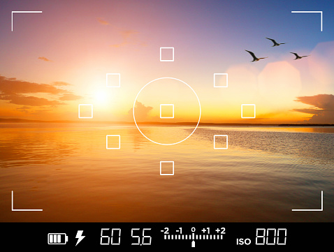 View through the viewfinder of a DSLR camera of photographer and traveler taking a picture of a beautiful sunset over the sea, with birds flying away.  Display shows data about exposure, shutter speed, aperture, ISO sensibility and focus points. Current settings are: 1/60s shutter speed, f:5.6 aperture, ISO 800 film sensibility. Icons show also battery charge, flash availability and exposure compensation levels.