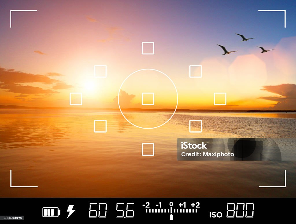 View through camera viewfinder, photographing a sunset View through the viewfinder of a DSLR camera of photographer and traveler taking a picture of a beautiful sunset over the sea, with birds flying away.  Display shows data about exposure, shutter speed, aperture, ISO sensibility and focus points. Current settings are: 1/60s shutter speed, f:5.6 aperture, ISO 800 film sensibility. Icons show also battery charge, flash availability and exposure compensation levels. Speed Stock Photo