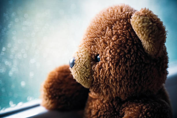 Vintage teddy bear looking through  wet window. Portrait of a toy bear by a sunlit window with rain drops on it. After the rain comes the Sun concept. Loneliness.