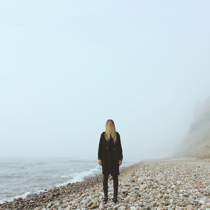 Young hipster female in black coat on the beach in a cold foggy day.