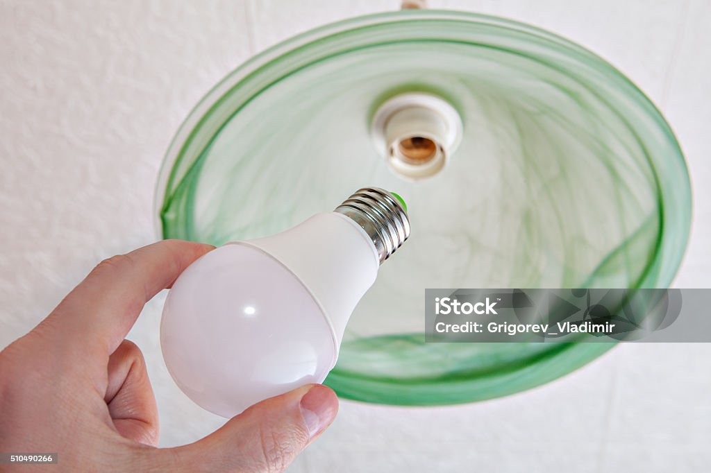 Close-up of energy-saving LED light bulb in human hand. Hand holding an energy-efficient LED lamp, replacement of the light bulb in the ceiling fixture with a shade of green frosted glass, close-up. Change Stock Photo