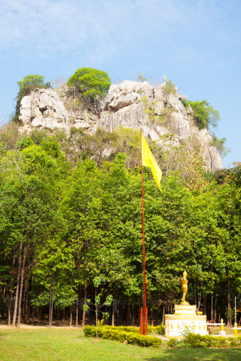 Grounds of temple aroundand at rock in small village Paragmee on province of Phitsanulok. At right side is small golden buddha statue, flag behind. In top of rock are more flags. Countryside temple with temple building and old cave temple inside of rock.