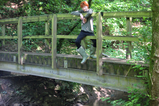Photo showing a young teenage boy with short red hair, pictured sitting and leaning on the railings of a wooden bridge, in the dappled shade of a woodland, looking down at the stream beneath him.  The forest canopy is lit by the afternoon sun behind them, causing a blurred summer background of green leaves.