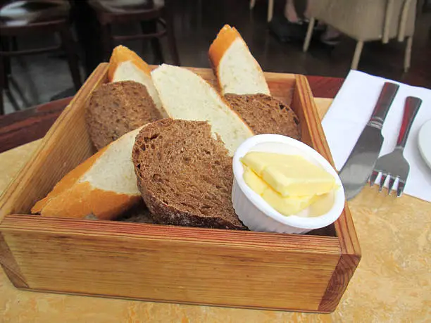 Photo showing a batch a freshly baked homemade bread / French sticks, both wholemeal and white bread, that have been sliced into chunks and are pictured being served in a wooden box / basket, with a small dish of butter on the side.