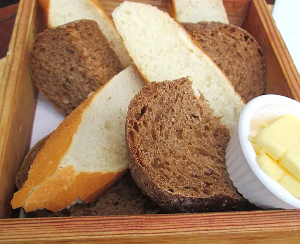 Photo showing a batch a freshly baked homemade bread / French sticks and loaves, both wholemeal and white bread, that have been sliced into chunks and are pictured being served in a wooden box / basket, with a small dish of butter on the side.