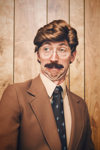 A 1970's - 1980's style newscaster TV reporter or business man stands in front of a wood paneled wall with an ugly expression on his face.  Styling is very stereotypical of that era; over the top side parted hair, large mustache, and oversized glasses are complimented with brown clothes and background.  Vertical image.