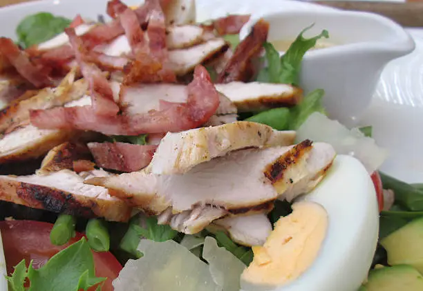 Photo of a Paysanne chicken and bacon salad, served in a restaurant on a white plate.  This meal consisted of grilled and sliced chicken breast, thick bacon / gammon ham, sliced boiled eggs, chopped tomatoes, a salad of green lettuce leaves, avocado chunks, French beans / green beans and grated Parmesan cheese / Parmesan shavings.