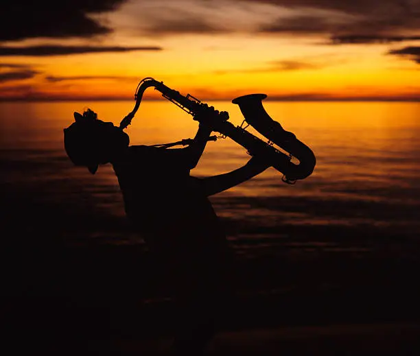 A young teen boy wearing a funky hat plays his saxophone with gusto on the beach at sunset.  The young musician is silhouetted against the yellow sky with the setting sun positioned over the end of the saxophone.  Scanned 6x7 film.  Copy space and room for cropping to desired size.