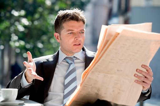 young attractive businessman sitting outdoors having coffee cup for breakfast reading newspaper news upset angry and worried with surprised shocked face expression in business concept