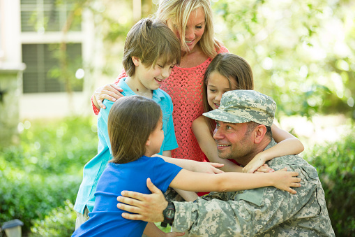 Handsome U.S.A. military soldier being welcomed home by his family.  Wife, two little girls and one boy children. Home in background. He wears his military uniform and is excited to see his family.  The children and wife eagerly give him a big hug. Thank you veterans for your service to your country!  Memorial Day, Veteran's Day.