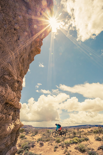 a woman pedals past a rock formation with the sunshine lens flare underneath a cloud filled blue sky.  vertical wide angle composition taken in gallup, new mexico.