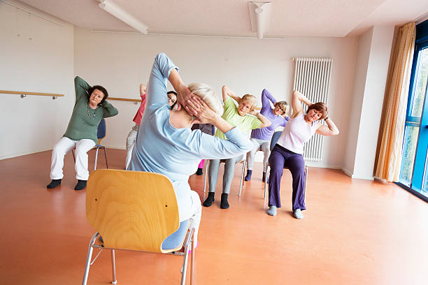 teacher and active senior women yoga class on chairs senior women exercising yoga and pilates sitting on chairs, following the instruction of their teacer community center photos stock pictures, royalty-free photos & images